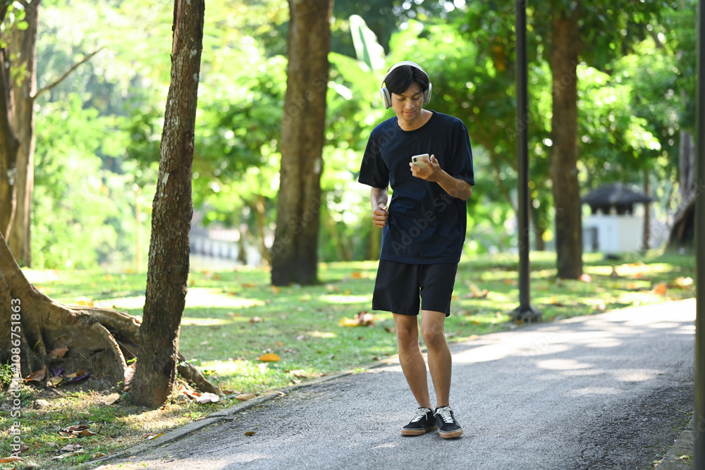 Young man holding mobile phone jogging along a paved path in a park on a sunny day Young man holding mobile phone jogging along a paved path in a park on a sunny day