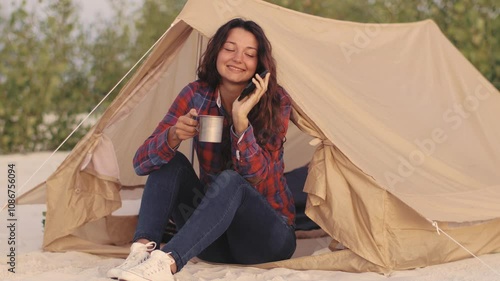 A woman sits outside her tent, happily talking on her phone while holding a coffee mug during a serene camping morning. Perfect for outdoor, travel, and lifestyle content focused on nature and