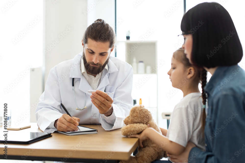 Caucasian male doctor examines temperature of young Caucasian girl with mother by her side. Doctor records information in notebook during appointment. Caucasian male doctor examines temperature of young Caucasian girl with mother by her side. Doctor records information in notebook during appointment.