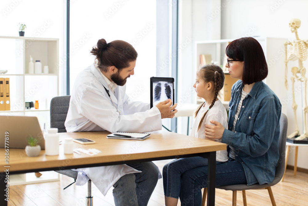 Caucasian male doctor discusses lung x-ray with young Caucasian girl and her mother in clinic. Doctor wearing white coat, using tablet to show medical image while girl attentively listens. Caucasian male doctor discusses lung x-ray with young Caucasian girl and her mother in clinic. Doctor wearing white coat, using tablet to show medical image while girl attentively listens.