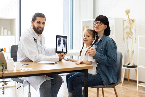 Caucasian male doctor shows lung x-ray to young girl and mother in bright clinic setting. Doctor wears lab coat and uses tablet for demonstration. Girl appears attentive and mother sits supportively.