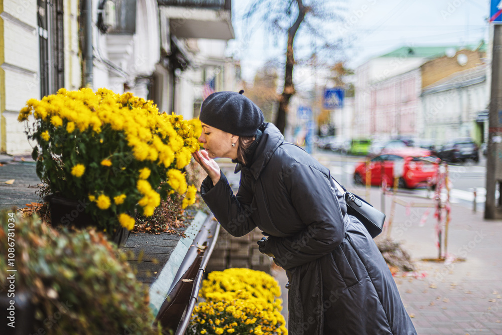 A woman on an autumn city street near a flower shop is interested in yellow flowers. The woman is wearing a black coat and beret. A woman on an autumn city street near a flower shop is interested in yellow flowers. The woman is wearing a black coat and beret.