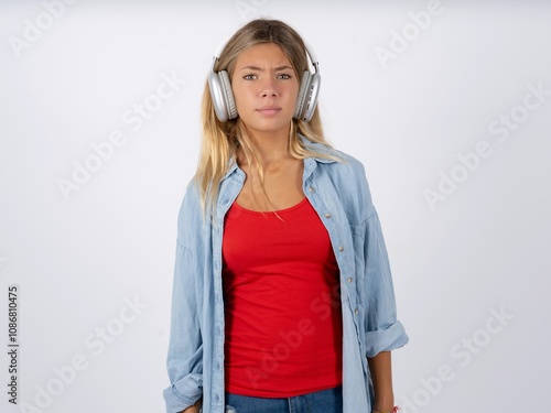 Serious displeased Beautiful teen girl wearing denim jacket  looks puzzled at camera being angry wears stereo headphones listens music while walking at street photo