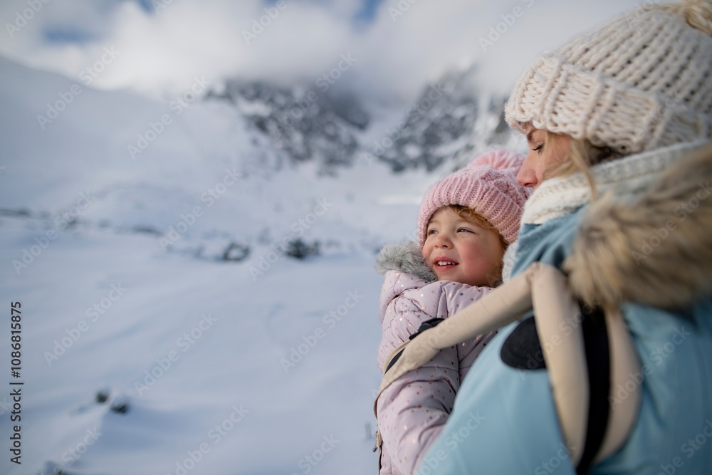 Obraz premium Mother standing in the middle of snowy landscape with daughter in baby carrier. Mom and girl enjoying winter holiday in the mountains.