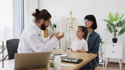 Caucasian male doctor offers medicine bottle and blister with pills to young daughter and mother in clinic. Little girl sits on mother's lap, interacting with medical professional.