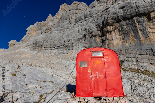 Alpine bivaco mountain hut shelter located in rugged alpine terrain. Red bivacco shelter in Dolomites. Compact structure against stunning landscapes, surrounded by peaks, valleys, and nature. photo