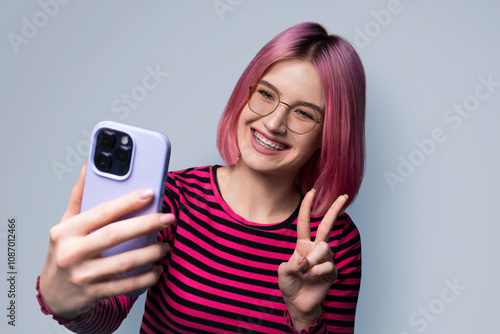 Portrait photo - pink hair woman in braces, tooth brackets wear eye glasses making taking selfie or show her viewer positive result with cell phone smartphone, v-sign gesture isolated grey background photo