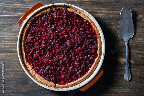 Delicious autumn pie on shortcrust pastry with cranberries on a wooden table with a cake spoon. Cranberry cake with jam and sugar in a baking dish, rustic style, closeup, top view