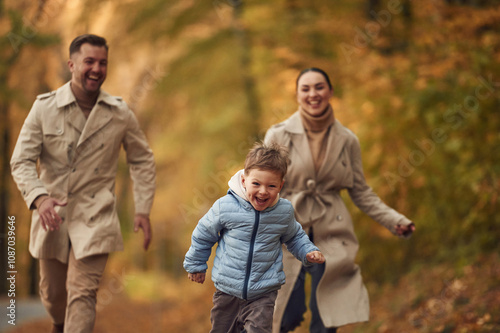 Boy is running forward. Happy family are together in the autumn forest near the road