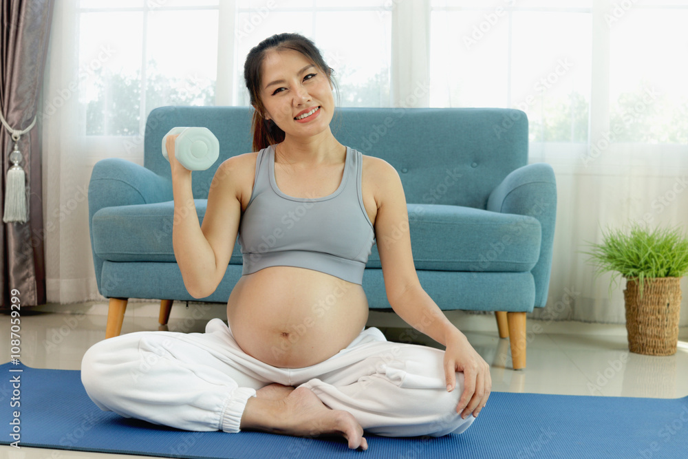 Asian pregnant woman sits on yoga mat on living room floor, do exercises at home. Beautiful pregnancy mother with big belly lifting dumbbell up workout to health and physical ability. Happy family Asian pregnant woman sits on yoga mat on living room floor, do exercises at home. Beautiful pregnancy mother with big belly lifting dumbbell up workout to health and physical ability. Happy family