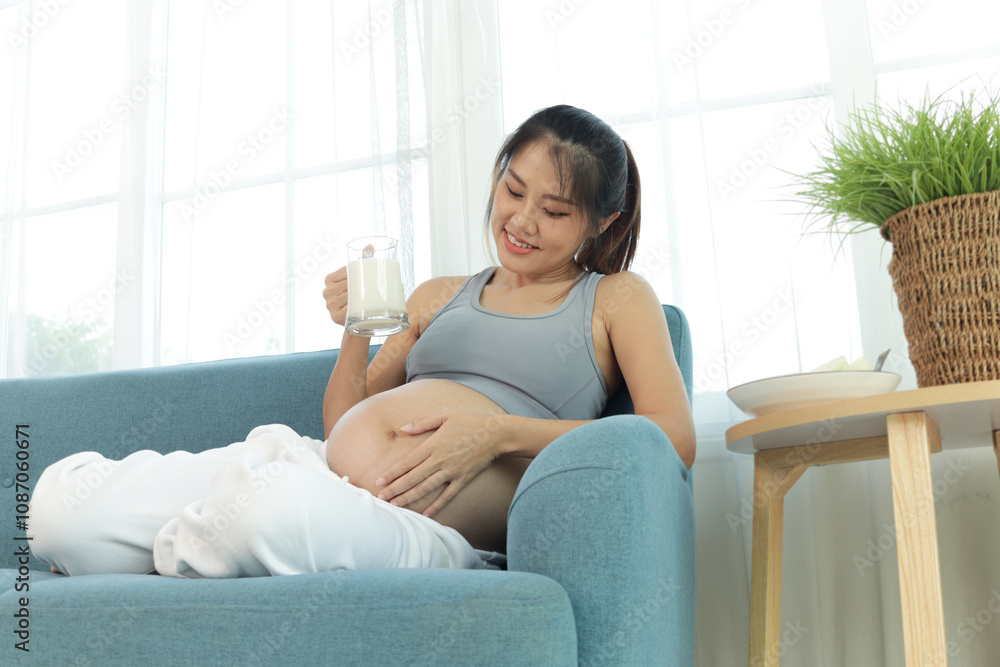 Happy Asian pregnant woman drinking milk from glass while sitting on sofa couch at living room. Beautiful pregnancy mother with big tummy drinking rich calcium food for her baby. Happy family at home Happy Asian pregnant woman drinking milk from glass while sitting on sofa couch at living room. Beautiful pregnancy mother with big tummy drinking rich calcium food for her baby. Happy family at home