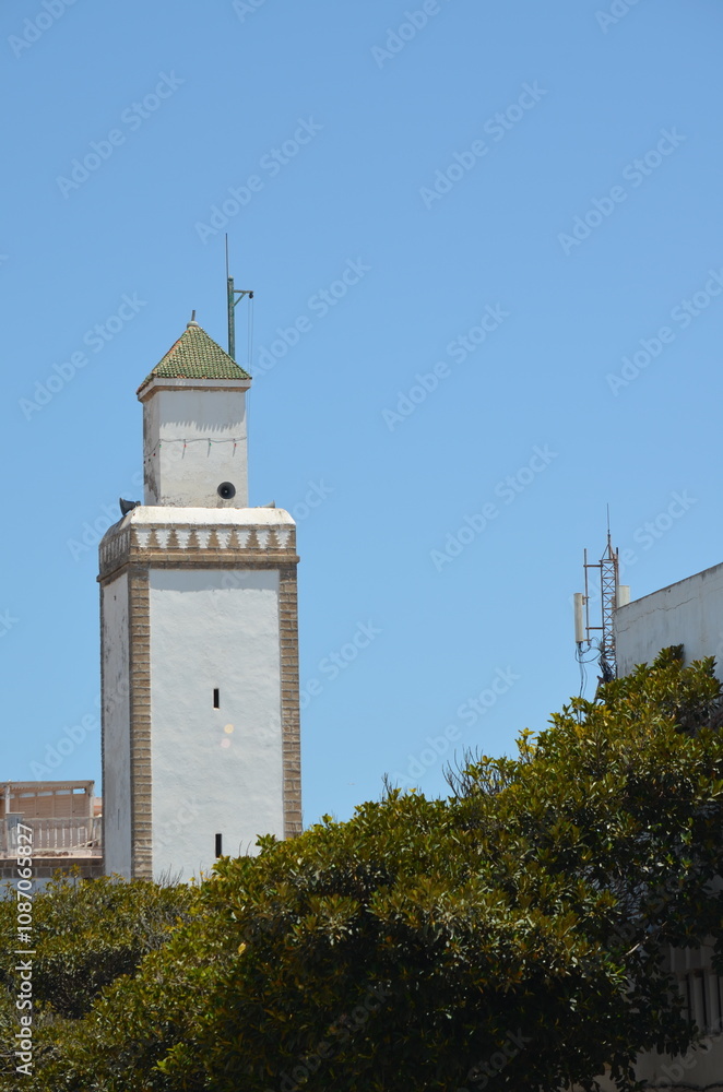 Tall white minaret with a green-tiled roof standing against a clear blue sky in Essaouira, surrounded by lush greenery, blending tradition with the coastal landscape
Tall white minaret with a green-tiled roof standing against a clear blue sky in Essaouira, surrounded by lush greenery, blending tradition with the coastal landscape