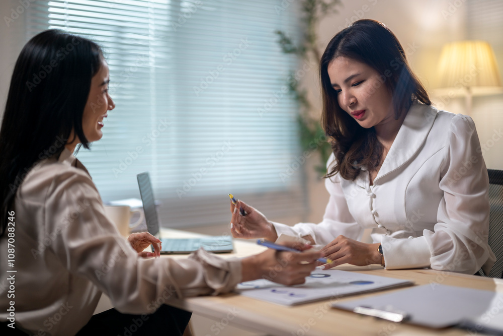 Two women are sitting at a desk, one of them holding a pen Two women are sitting at a desk, one of them holding a pen