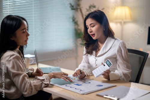 Two women are sitting at a desk, one of them holding a calculator