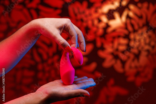 In soft neon light a woman showcases a small pink vibrator designed for intimate pleasure. The glowing red backdrop adds a touch of elegance to this intimate moment.