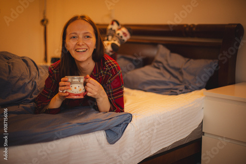 Morning bliss: A young woman in festive pajamas enjoys a cup of tea in bed, radiating happiness and warmth in a cozy bedroom setting. Perfect for themes of self-care, relaxation photo