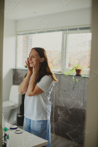 Happy Woman Applying Face Cream Moisturizing And Caring For Skin Smiling To Her Reflection In Mirror Standing In Modern Bathroom At Home. Facial Skincare And Pampering. Selective Focus photo