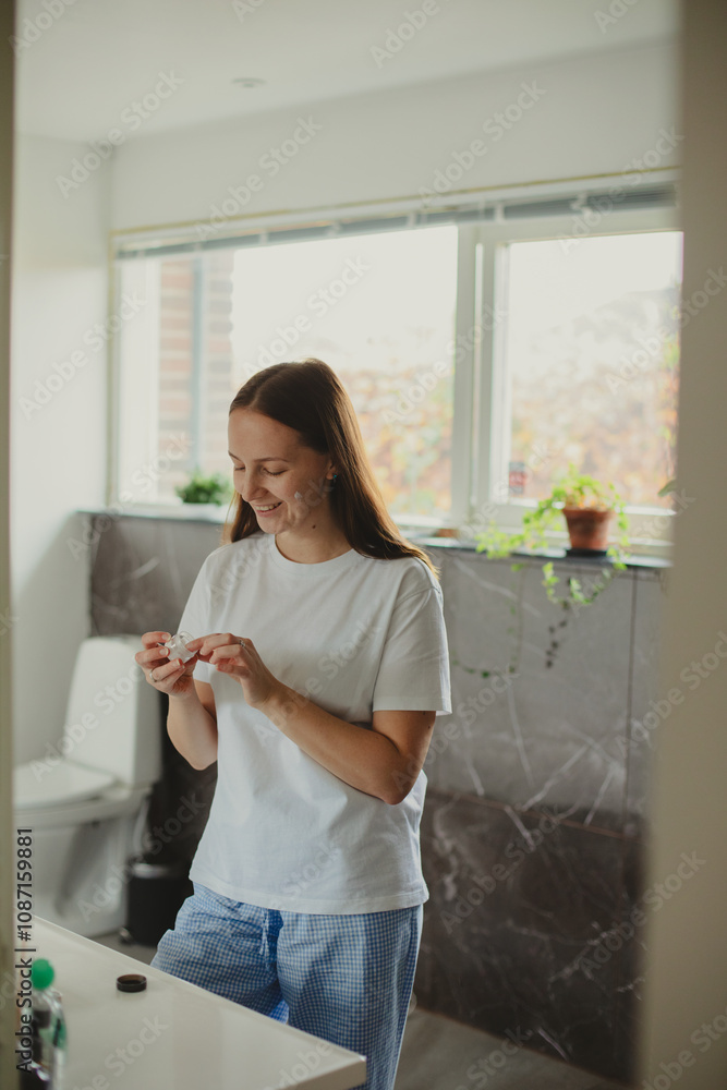 Happy Woman Applying Face Cream Moisturizing And Caring For Skin Smiling To Her Reflection In Mirror Standing In Modern Bathroom At Home. Facial Skincare And Pampering. Selective Focus Happy Woman Applying Face Cream Moisturizing And Caring For Skin Smiling To Her Reflection In Mirror Standing In Modern Bathroom At Home. Facial Skincare And Pampering. Selective Focus