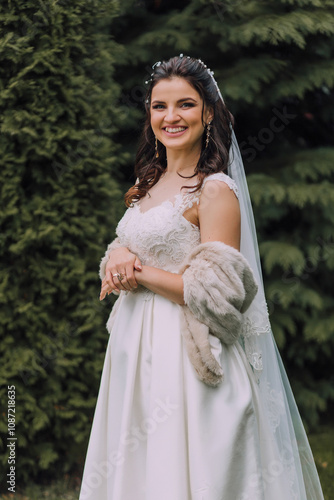 A woman in a white dress is smiling and holding her hands together. She is wearing a fur stole and a veil photo