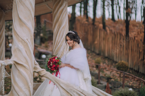 A bride is standing on a stone staircase with a bouquet of red flowers in her hand. She is wearing a white dress and a white fur stole. The scene is set in a forest, with trees in the background photo