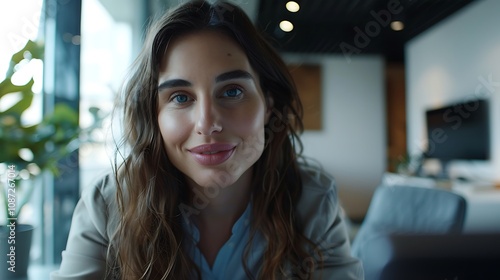 A woman with wavy hair smiles warmly at the camera in a modern office setting.