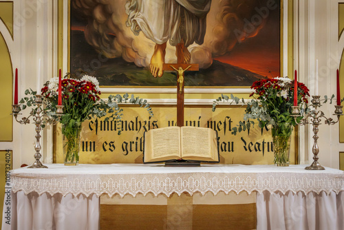 A church altar featuring an open Bible, a wooden crucifix, and floral arrangements on either side. The background includes a religious painting and decorative script