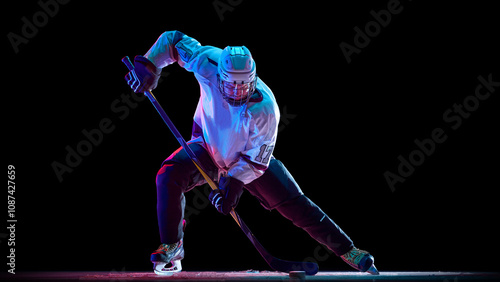 Photography Determined athlete, hockey player in white uniform in motion during game, controlling puck with stick against black studio background in neon light