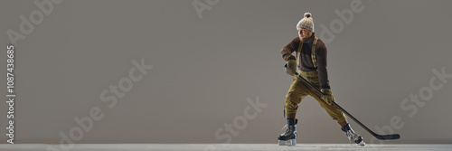 Photography Barded man, hockey player wearing sweater, hat, pants with suspenders, posing with stick against grey background