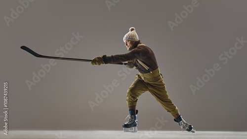 Canvas Print Bearded man, hockey player in retro outfit, wearing knitted hat and sweater, pants with suspenders, in motion with stick on studio background