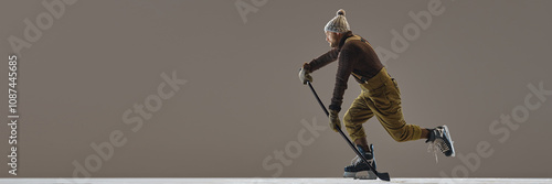 Photography Barded man, hockey player wearing sweater, hat, pants with suspenders, in motion with stick against grey background