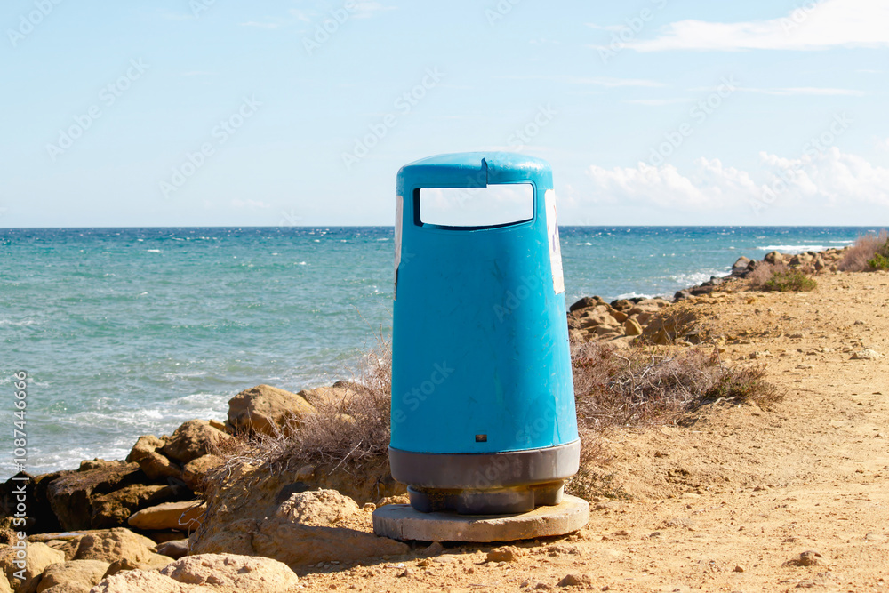 A vibrant blue trash bin set against a rocky coastal pathway and the vast ocean in the background. A vibrant blue trash bin set against a rocky coastal pathway and the vast ocean in the background.
