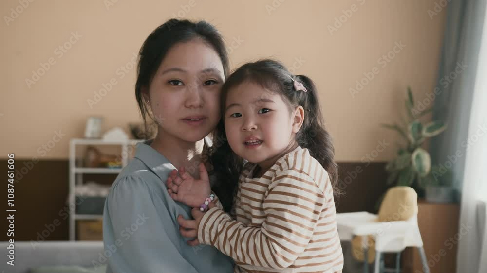 Young Asian woman in blue shirt holding adorable little daughter on hands while standing in the center of spacious living room at home and both looking at camera