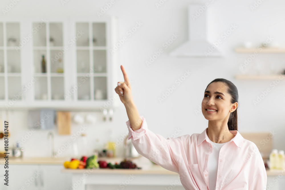 Smart Home Interface Concept. Portrait Of Positive Cheerful Casual Young Female Pointing Finger Up In The Air Pushing Button On Invisible Screen Standing In Kitchen Interior In Her New Apartment Smart Home Interface Concept. Portrait Of Positive Cheerful Casual Young Female Pointing Finger Up In The Air Pushing Button On Invisible Screen Standing In Kitchen Interior In Her New Apartment