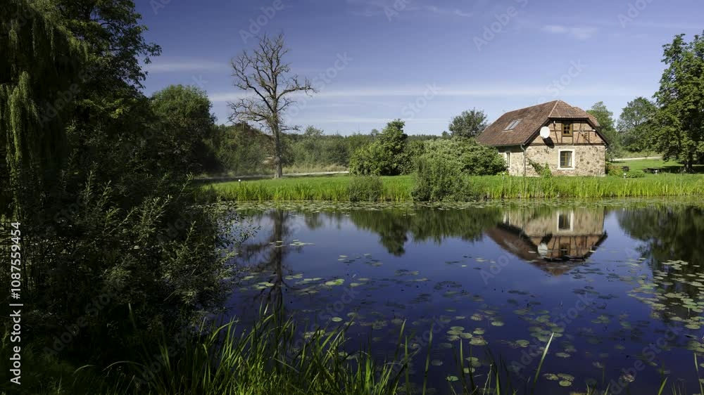 Rural two-story stone house among the trees near the pond. Lilies float on the water against the backdrop of reflections. Green foliage on a sunny summer day. Jaunmoku Castle. Latvia Rural two-story stone house among the trees near the pond. Lilies float on the water against the backdrop of reflections. Green foliage on a sunny summer day. Jaunmoku Castle. Latvia