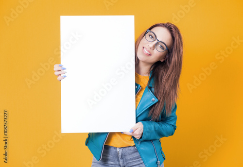Smiling young women holding white board, wearing blue leather jacket, isolated on yellow background. One cute girl showing blank empty paper billboard with blank space for text
