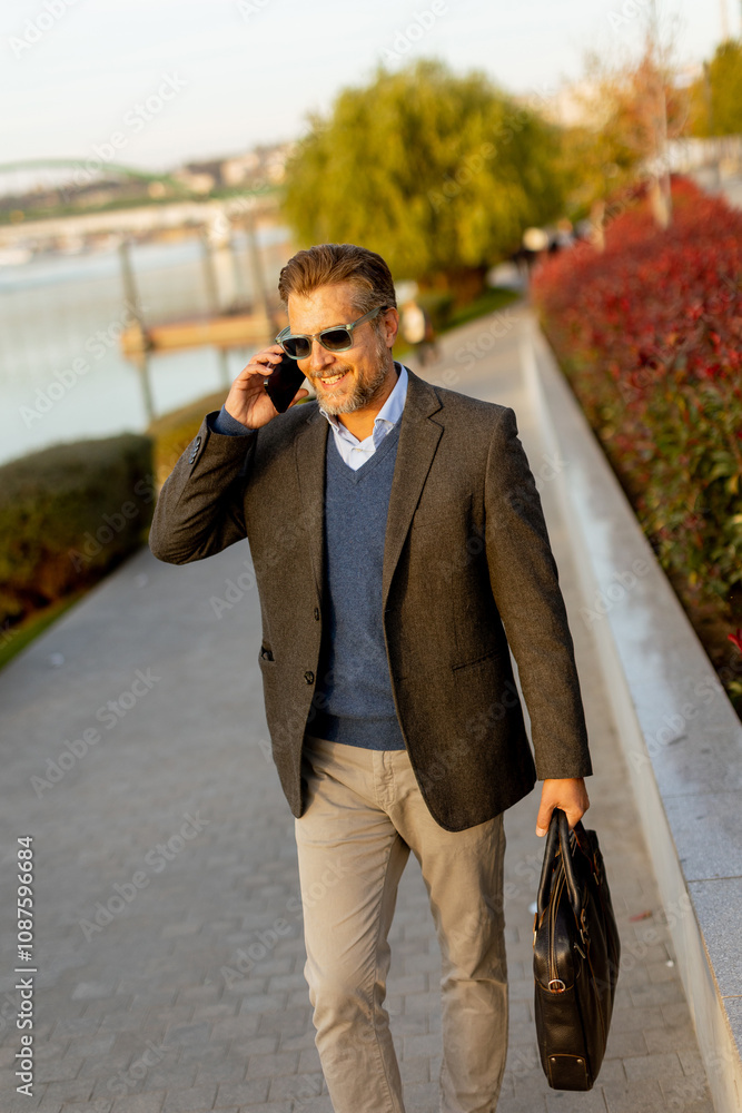 A stylish man enjoys a leisurely stroll along the waterfront, chatting on his phone amidst autumn foliage in the golden afternoon light A stylish man enjoys a leisurely stroll along the waterfront, chatting on his phone amidst autumn foliage in the golden afternoon light