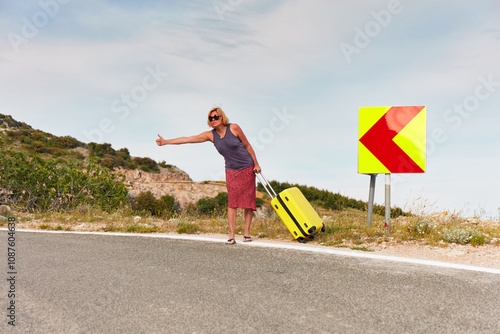 Attractive mature woman with a large yellow suitcase stopping hitchhiking on the road in southern Europe. Summer evening, slightly cloudy sky.