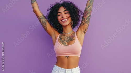 Joyful woman with curly hair and tattoos posing against a purple background d...