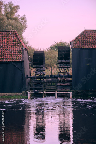 Historic watermills of Collse Molen in Eindhoven, Netherlands, surrounded by calm water, trees, and a soft pink sky during sunset. photo