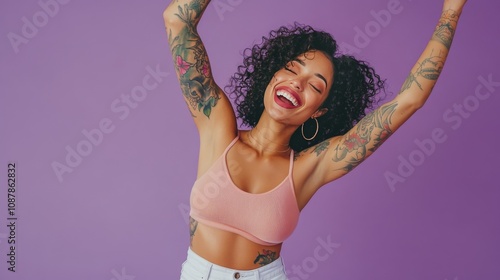 Joyful woman with curly hair and tattoos posing against a purple background d...