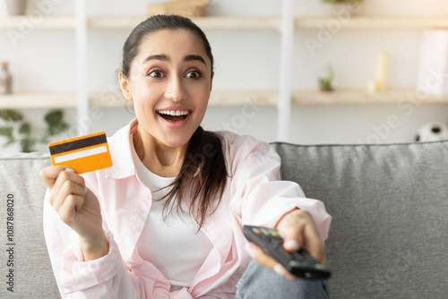 Wow. Portrait of amazed emotional young woman sitting on comfortable sofa in living room, using tv remote controll and holding yellow golden credit debit card, selective focus photo