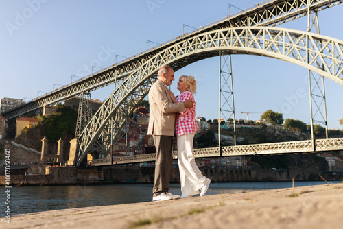 A couple embraces warmly by the riverbank, enjoying the serene view beneath a striking bridge in Porto during a sunny afternoon. Their joyful interaction radiates love and companionship.