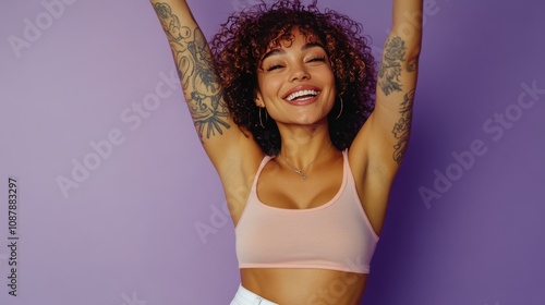 Joyful woman with curly hair and tattoos posing against a purple background d...