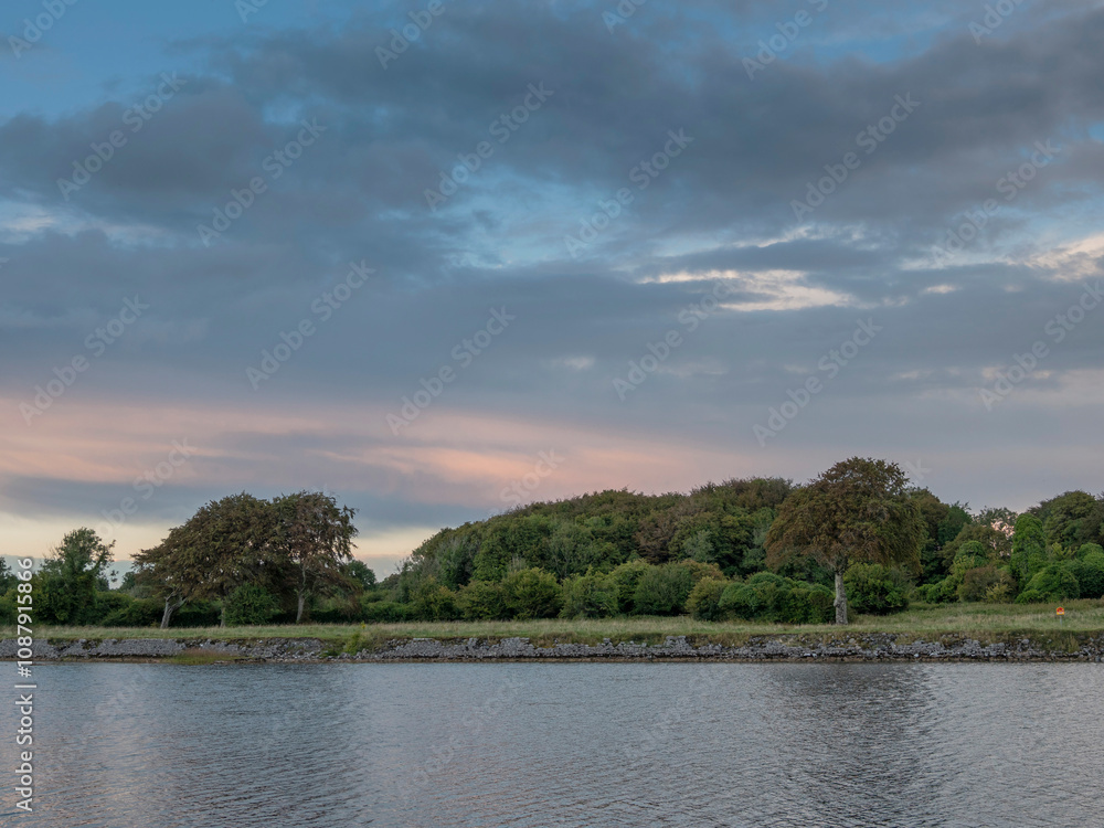 A calm lake with trees in the background. The sky is cloudy and the sun is setting. Blue hour after sunset. Simple nature background. A calm lake with trees in the background. The sky is cloudy and the sun is setting. Blue hour after sunset. Simple nature background.