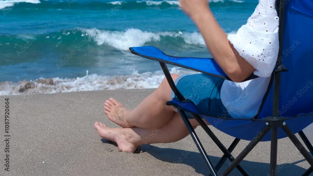 Relaxing in chair on beach enjoying ocean waves. Woman wearing a white shirt and blue jean shorts sits in a blue beach chair on a sunny beach, resting and enjoying the cool water washing over her feet Relaxing in chair on beach enjoying ocean waves. Woman wearing a white shirt and blue jean shorts sits in a blue beach chair on a sunny beach, resting and enjoying the cool water washing over her feet