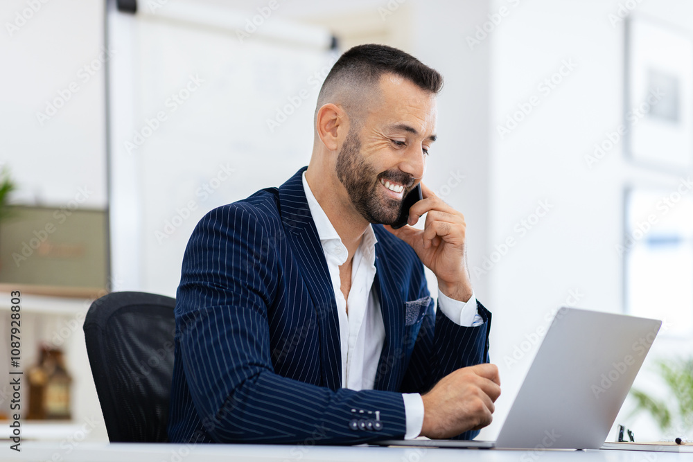 A well-dressed man smiles as he has a phone conversation while working on his laptop in a bright, contemporary office environment. He appears focused and engaged. A well-dressed man smiles as he has a phone conversation while working on his laptop in a bright, contemporary office environment. He appears focused and engaged.