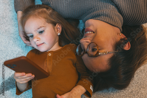 With her mother lying close by, a young girl intensely focuses on her smartphone, capturing a peaceful moment of exploration and learning in a domestic setting. photo