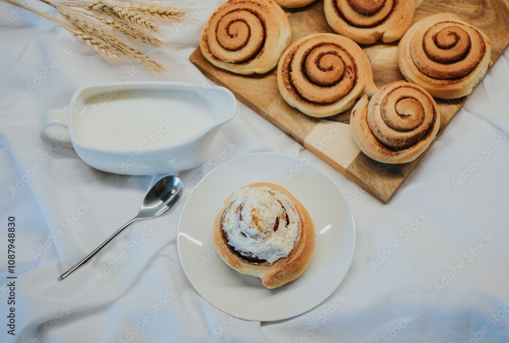 Fresh cinnabon on a plate on a background of other buns on a wooden tray and milk with a spoon Fresh cinnabon on a plate on a background of other buns on a wooden tray and milk with a spoon