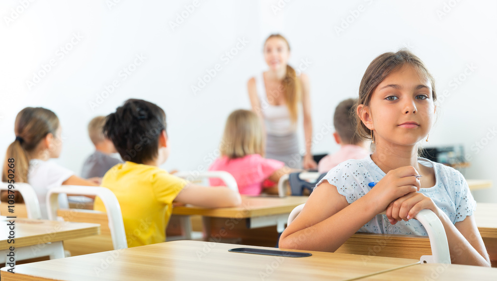 Smiling positive small school girl sitting at school desk in classroom on background with classmates Smiling positive small school girl sitting at school desk in classroom on background with classmates