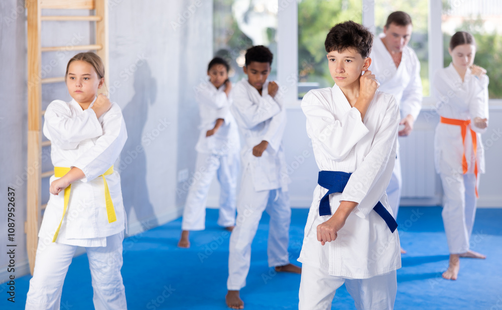 Determined boy in white kimono practicing punches in gym during martial arts workout with multiracial group of tweens. Shadow fight, combat sports training concept Determined boy in white kimono practicing punches in gym during martial arts workout with multiracial group of tweens. Shadow fight, combat sports training concept